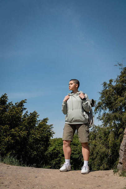 Junge, selbstbewusste, kurzhaarige Wanderin in lässiger Kleidung mit Rucksack und wegschauendem Blick im Hintergrund mit Bäumen und blauem Himmel, Übersetzung von Tätowierung: Liebe - Foto, Bild