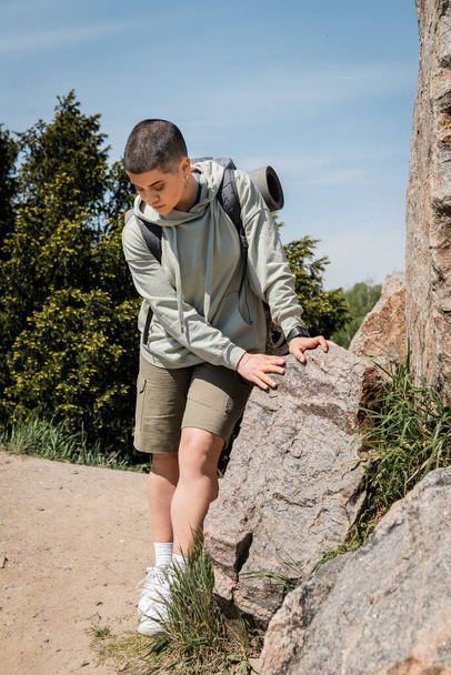 Young short haired female hiker in casual clothes with backpack looking at sneakers while standing near stones with trees and blue sky at background, trekking through rugged terrain - Fotó, kép