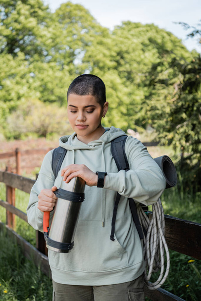 Young short haired female tourist with backpack and fitness tracker holding thermos while standing with blurred natural landscape at background, trekking through rugged terrain, summer - Photo, Image
