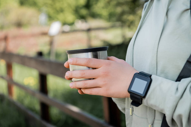 Cropped view of young woman tourist with fitness tracker holing cup of thermos and standing with blurred nature at background, finding serenity in nature, summer - Photo, Image