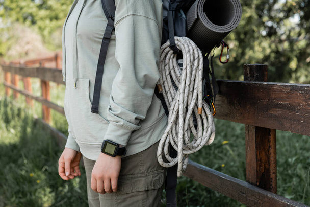 Ausgeschnittene Ansicht einer jungen Wanderin mit Fitness-Tracker, Rucksack und Reiseausrüstung mit verschwommener Landschaft im Hintergrund, die Gelassenheit in der Natur findet, Sommer - Foto, Bild