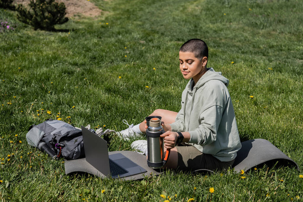Young short haired woman tourist holding thermos while sitting on fitness mat near laptop and backpack on grassy lawn with flowers, finding serenity in nature, summer, digital nomad  - Photo, Image