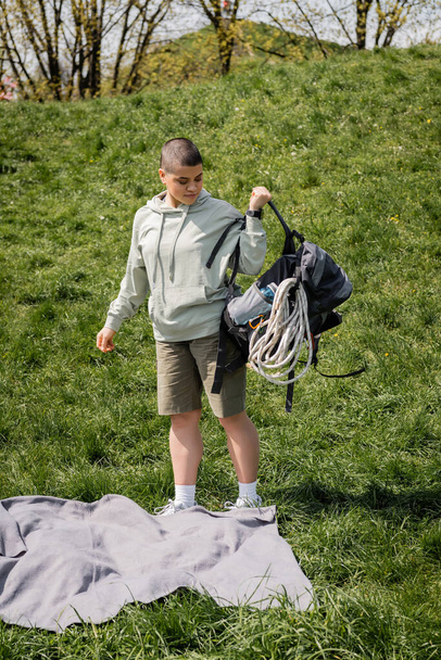 Young short haired female hiker with fitness tracker holding backpack with travel equipment while standing near blanket on grassy lawn with hill at background, connecting with nature concept - Zdjęcie, obraz