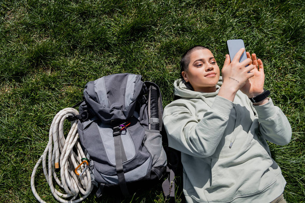Top view of young short haired and tattooed female traveler in casual clothes using smartphone while lying near backpack on grassy lawn, solo hiking journey concept , summer - Photo, Image