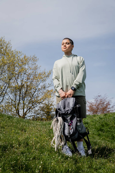 Mladá krátkosrstá turistka s chytrou batohem s cestovním vybavením a pohledem stranou, stojící na travnatém kopci, projíždějící malebnou krajinou, léto - Fotografie, Obrázek