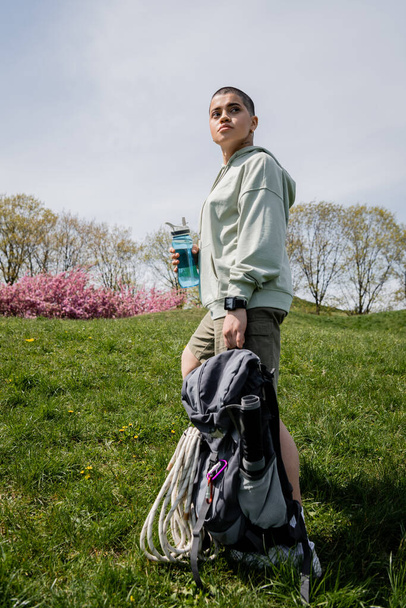 Krótkowłosa podróżniczka ze smartwatchem trzymająca plecak ze sprzętem podróżniczym i butelką sportową, odwracając wzrok na trawiastej łące w tle, przemierzając malowniczy krajobraz - Zdjęcie, obraz