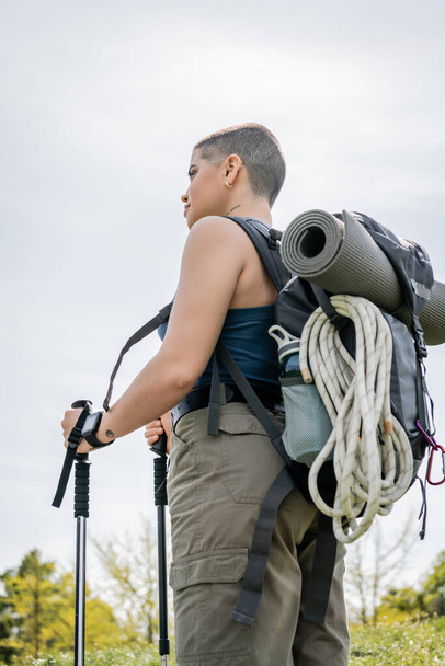 Niski kąt widzenia młodych krótkowłosych i wytatuowanych kobiet wędrowca ze smartwatch i plecakiem trzymając drągi trekkingowe i patrząc z natury na tle, samotne wędrówki koncepcja podróży  - Zdjęcie, obraz