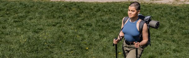Hochwinkelaufnahme einer jungen kurzhaarigen Backpackerin mit Rucksack, die lächelt und Trekkingstöcke hält, während sie im Hintergrund auf einer grasbewachsenen Wiese spaziert, Einzelwanderungskonzept, Banner - Foto, Bild