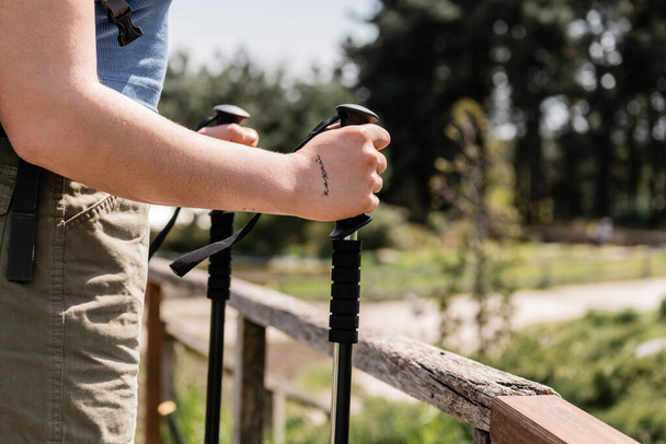 Cropped view of young tattooed woman tourist holding trekking poles while standing near wooden fence with nature at background, reconnecting with yourself in nature, Translation of tattoo: love - Φωτογραφία, εικόνα