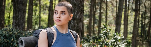 Young short haired female traveler with backpack and fitness mat looking away while standing in blurred green forest, reconnecting with yourself in nature concept, banner  - Photo, Image