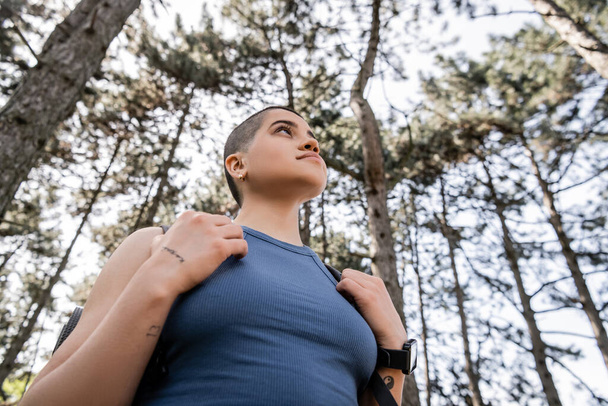 Low angle view of young short haired and tattooed female hiker with smartwatch and backpack looking away while standing in blurred summer forest, wanderlust vibes concept - Fotografie, Obrázek