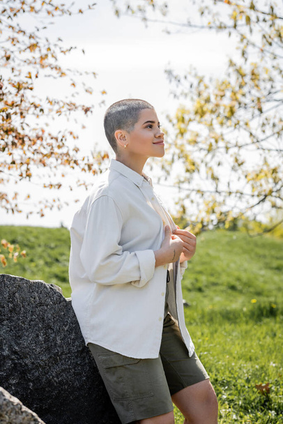 Positive young short haired female hiker wearing casual shirt while looking away and standing near stones with blurred scenic landscape at background, Translation of tattoo: love - Фото, зображення
