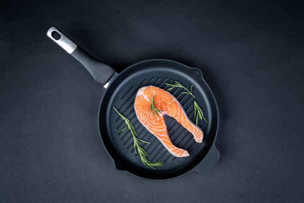 Fresh raw salmon slice with rosemary arranged in a grill pan on black background. Preparing healthy seafood dinner. - Φωτογραφία, εικόνα