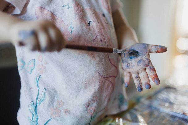 Close up of little girl painting on her own hand with tempera paint, using a paintbrush. - Zdjęcie, obraz