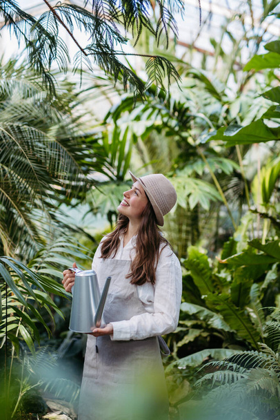 Portrait of beautiful female gardener taking care of plants in botanical garden. Botanist, horticulturist watering plants with watering can. - Фото, изображение
