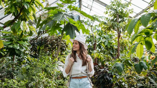 Portrait of a young woman with hat and backpack in botanical garden. Botanist in greenhouse. - Фото, изображение