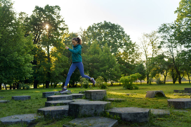 Full lenght portrait of beautiful fitness woman jumping on concrete obstacles in the park. Training for obstacle course, adventure racing. Workout at the park. - Φωτογραφία, εικόνα