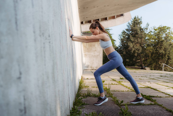 Side view of young sporty woman in sportswear excercising outdoors. Fitness woman stretching against concrete wall in the city. Healthy lifestyle concept. Street workout. - Foto, afbeelding
