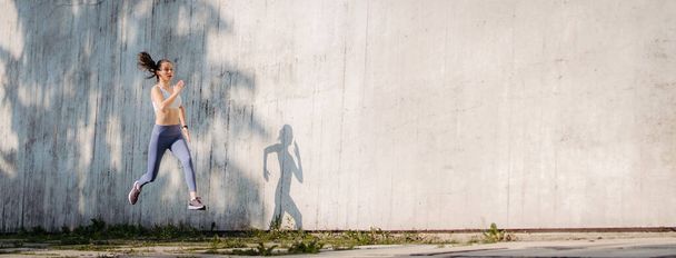 Female athlete in sportswear exercising in the city. Running in front of concrete wall casting shadow. Banner with copy space. - Φωτογραφία, εικόνα