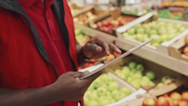 Comerciante masculino afroamericano del supermercado que usa uniforme rojo usando tableta digital mientras trabaja en el departamento de comestibles - Imágenes, Vídeo
