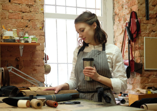 vrouw werkt in een zak maken studio, snijdt details en drink koffie - Foto, afbeelding