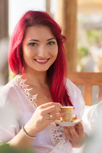 egy gyönyörű lány divatos vörös hajjal egy reggeli köntösben egy nyári teraszon a növények között nevet és kávézik.nő pihen. reggeli kávé. - Fotó, kép
