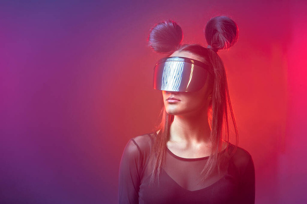 Porträt der schönen Cyber-Model Frau posiert mit futuristischer Brille auf dem Kopf und Neonlicht in einer virtuellen Tech-Umgebung - Foto, Bild
