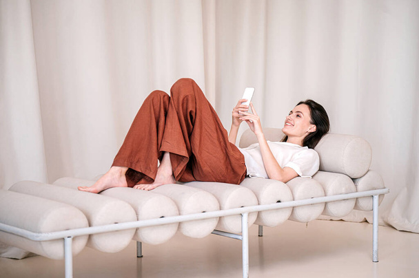 Junge Frau auf bequemem Hocker liegend, Smartphone in der Hand. Weibchen ruhen auf dem Bett im Zimmer, SMS per Handy. Mädchen auf weicher Couch mit Handy und Online-App zum Chatten - Foto, Bild