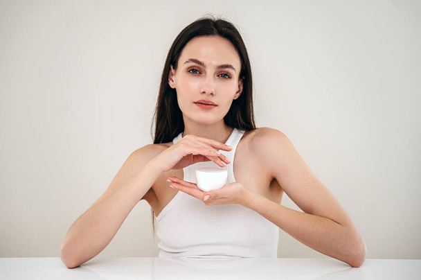 Concept of pampering beauty products for skincare and bodycare routine. Young woman with healthy hydrated skin holding jar with anti aging face cream, looking at camera - Valokuva, kuva