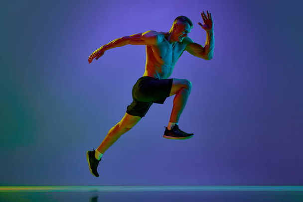 Dynamiczny obraz młodego człowieka z muskularnym, silnym, sprawnym ciałem, profesjonalnym biegaczem w ruchu na niebieskim tle pracowni w neonowym świetle. Pojęcie sportu, konkurencji, zdrowia, siły i szybkości, reklama - Zdjęcie, obraz