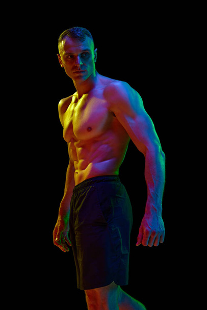 Pohledný muž s svalnatým, úlevou, fit, bez trička tělo pózovat proti černé studio pozadí v neonovém světle. Pojetí sportu, soutěže, péče o tělo, zdraví, síly, reklama - Fotografie, Obrázek