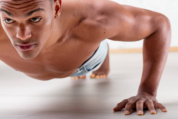 A black man performs wide-arm push-ups. With toned chest and arms out, this short-haired young man exercises shirtless on light wood flooring at home or gym. His strong arms and dedication to maintain - Zdjęcie, obraz