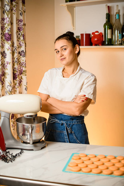 Woman chef stands near food processor. freshly baked halves of macaron dessert on silicone mat on blurred foreground. Homemade bakery concept - Photo, image