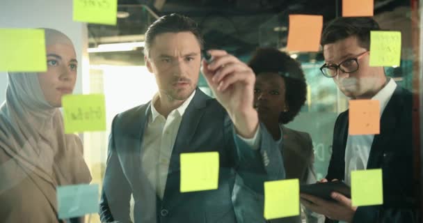 Multiethnic coworkers having business metting in office. Discusing about upcoming project. Creating goals and objectives from company. Handsome man writing on glass board where located stickers. - Footage, Video