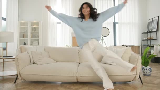 Щаслива жінка святкує день переїзду, насолоджуючись новою орендованою квартирою. Щастя веселі вихідні концепція повільний рух. Весела латинська позитивна жінка стрибає босоніж на дивані в сучасній вітальні вдома - Кадри, відео