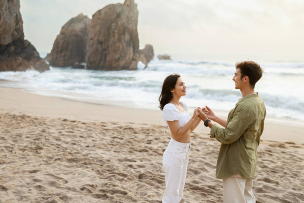 Milující pár drží za ruce a dívají se na sebe, mají romantické rande na pláži, stojí blízko pobřeží oceánu, volný prostor. Pár romantický moment na pobřeží - Fotografie, Obrázek
