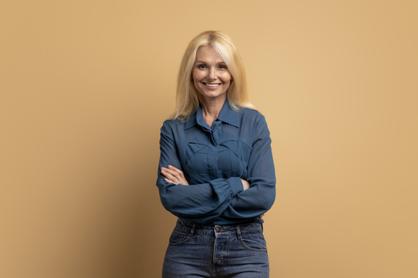 Stylish confident adult 50 years old attractive blonde woman standing arms crossed looking at camera at beige background. Portrait of sophisticated senior lady advertising products and services - Fotó, kép