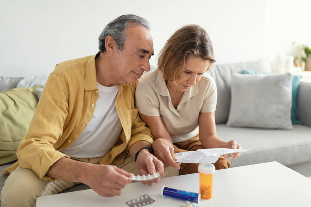 European aged spouses reading medications prescriptions for new pills or vitamins, sitting on couch in living room interior, free space. Nutritional supplement and healthcare concept - Φωτογραφία, εικόνα