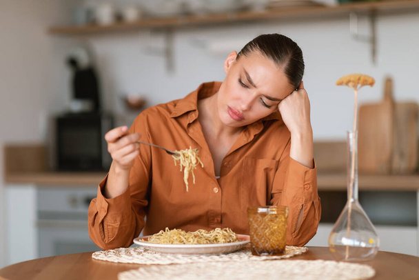 Young caucasian sad woman sitting at table in kitchen interior, looking at homemade pasta and dont want to eat, needs to keep diet - Zdjęcie, obraz