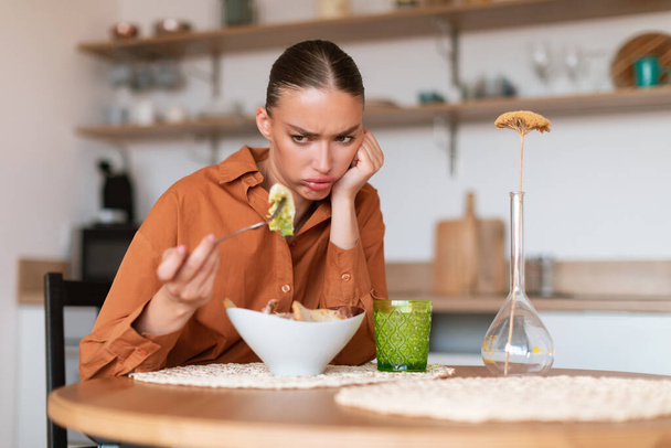 Taste trouble. Young caucasian woman showing dislike for her caesar salad, looking at it and dont want to eat, sitting at kitchen interior, free space - Photo, Image