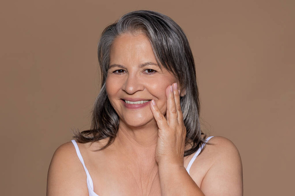 Smiling european senior gray haired woman enjoy perfect skin, good result after beauty therapy, isolated on beige studio background. Rejuvenation, spa treatment, lifting and beauty care - Photo, Image