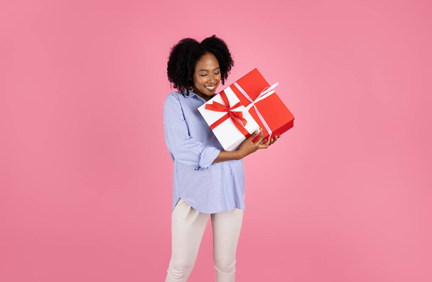 Счастливая африканская кудрявая беременная женщина тысячелетия в повседневной жизни с большим животом держит коробки с подарками, изолированные на розовом фоне студии. Подарок на праздничную вечеринку, ожидаем ребенка, день рождения - Фото, изображение