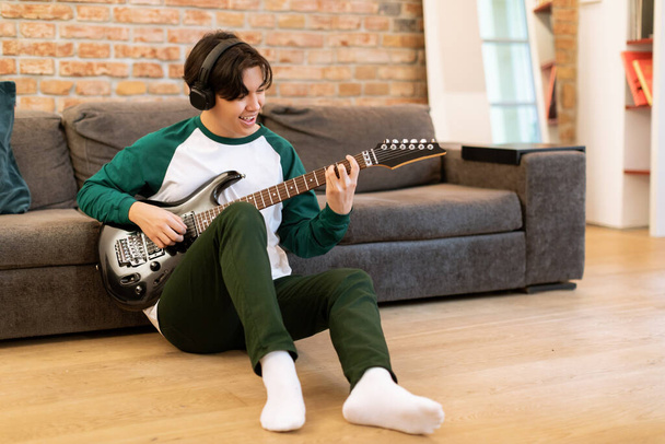 Edukacja muzyczna i wypoczynek. Młody japoński muzyk Guy Learning to Play, Praktykowanie gitara elektryczna siedzi na podłodze w nowoczesnym salonie wnętrz, noszenie słuchawki bezprzewodowe - Zdjęcie, obraz