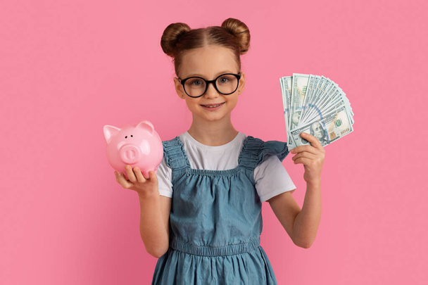 First Savings. Cute little girl holding dollar cash and piggy bank in hands, happy preteen female child in eyeglasses enjoying economy, recommending investment program, standing on pink background - Photo, Image