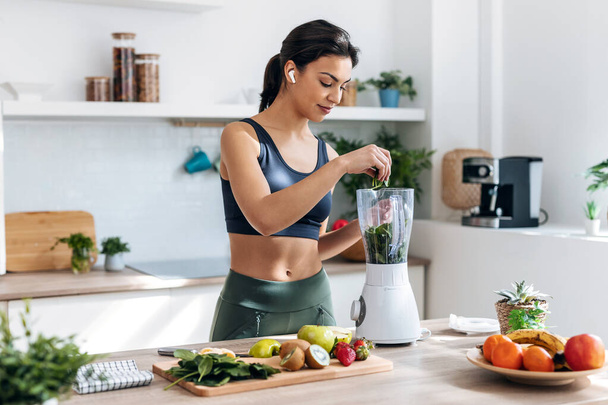 Shot of athletic woman preparing smothie with vegetables and fruits while listening music with earphones in the kitchen at home. - Foto, Imagem