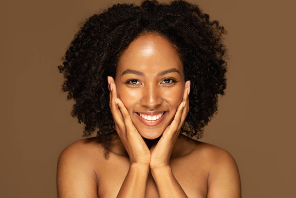 Nahaufnahme Studio-Foto von glücklichen schönen halbnackten jungen schwarzen Frau berührt ihr hübsches Gesicht mit beiden Händen und lächelt in die Kamera auf braunem Hintergrund, genießen ihre luxuriöse Hautpflege Routine - Foto, Bild