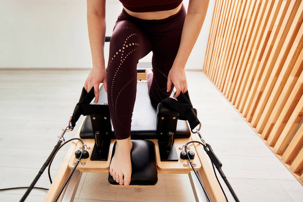 A young girl is doing Pilates on a reformer bed in a bright studio. A slender brunette in a burgundy bodysuit does exercises to strengthen the arms and shoulder girdle. Healthy lifestyle concept - Photo, Image
