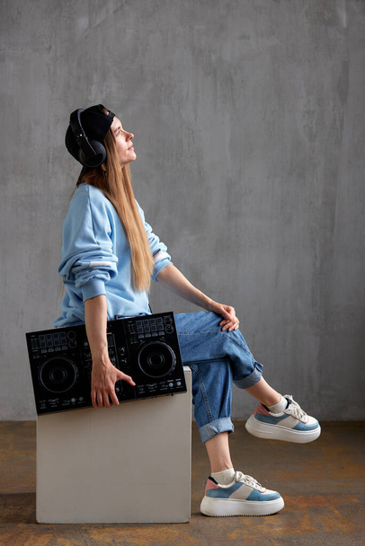 A young pretty long-haired DJ girl in a blue sweater, jeans, a black baseball cap and black headphones sits with a black mixing console in her hand. Studio shot, gray background - Fotó, kép