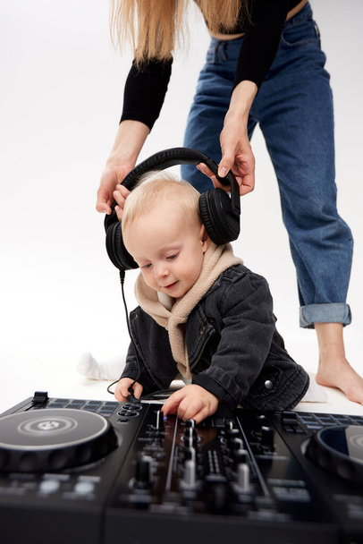 A cute kid in a beige hoodie and a funny black motorcycle jacket sits on the floor with a dj mixing console. A barefoot mom in blue jeans puts DJ headphones on his head. - Photo, image
