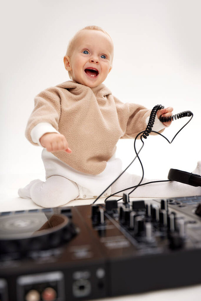 Słodki uśmiechnięty dzieciak w beżowej bluzie siedzi na podłodze i bawi się słuchawkami DJ-a i konsolą do miksowania DJ-a. Muzyka i zabawa. Izolacja na białym tle - Zdjęcie, obraz